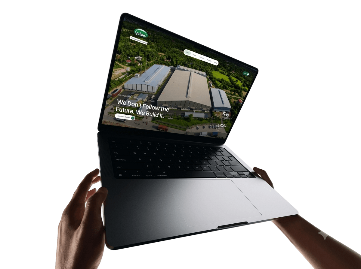 Hands holding open a laptop showing the PrimeNMS home page: an aerial photo of a steel warehouse under the line “We Don't Follow the Future. We Build It.”
