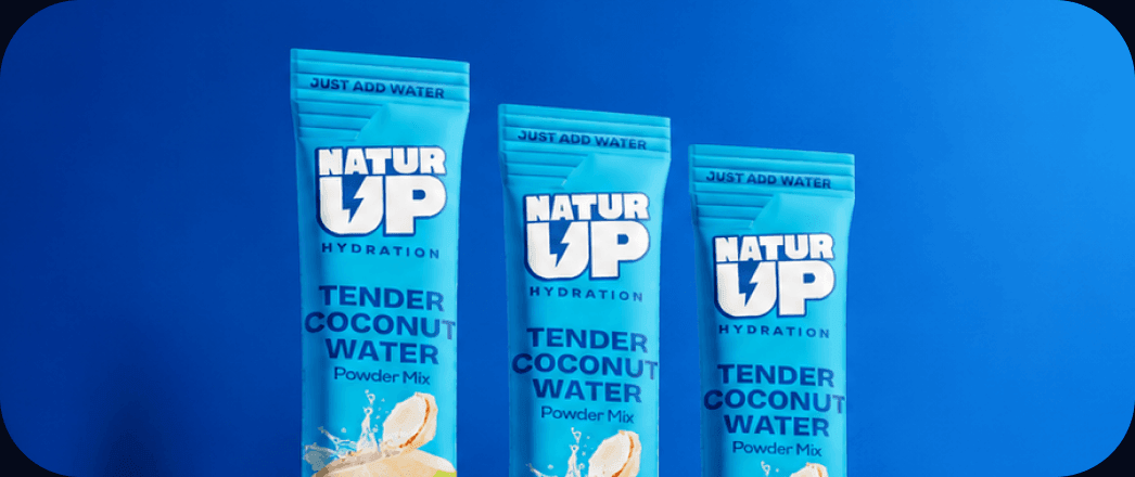 Three NaturUp tender coconut water sachets on a white shelf against a blue wall