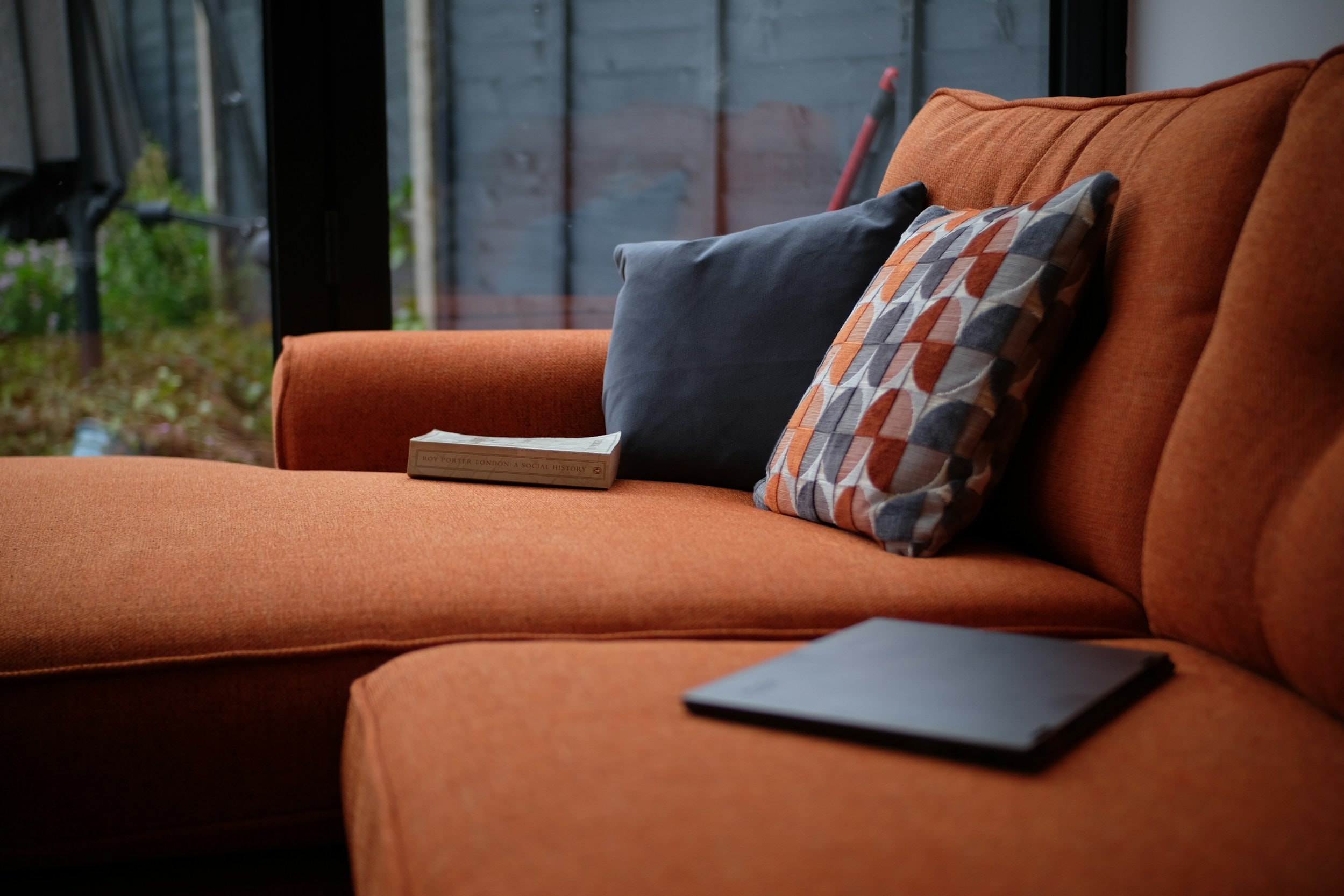 An orange sofa with patterned and charcoal cushions and a book, by a garden window