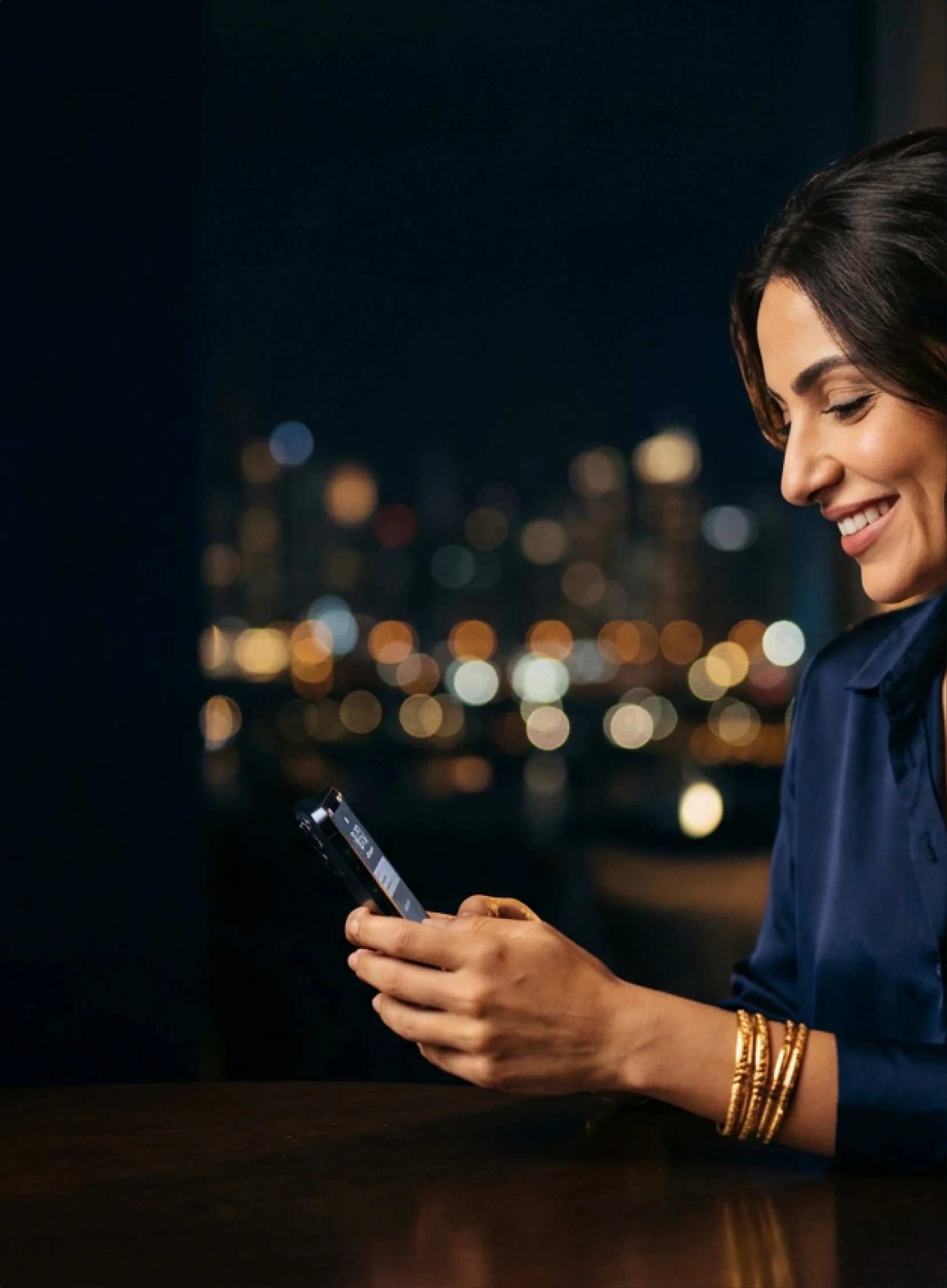 A woman in gold bangles smiling at her phone, city lights behind her