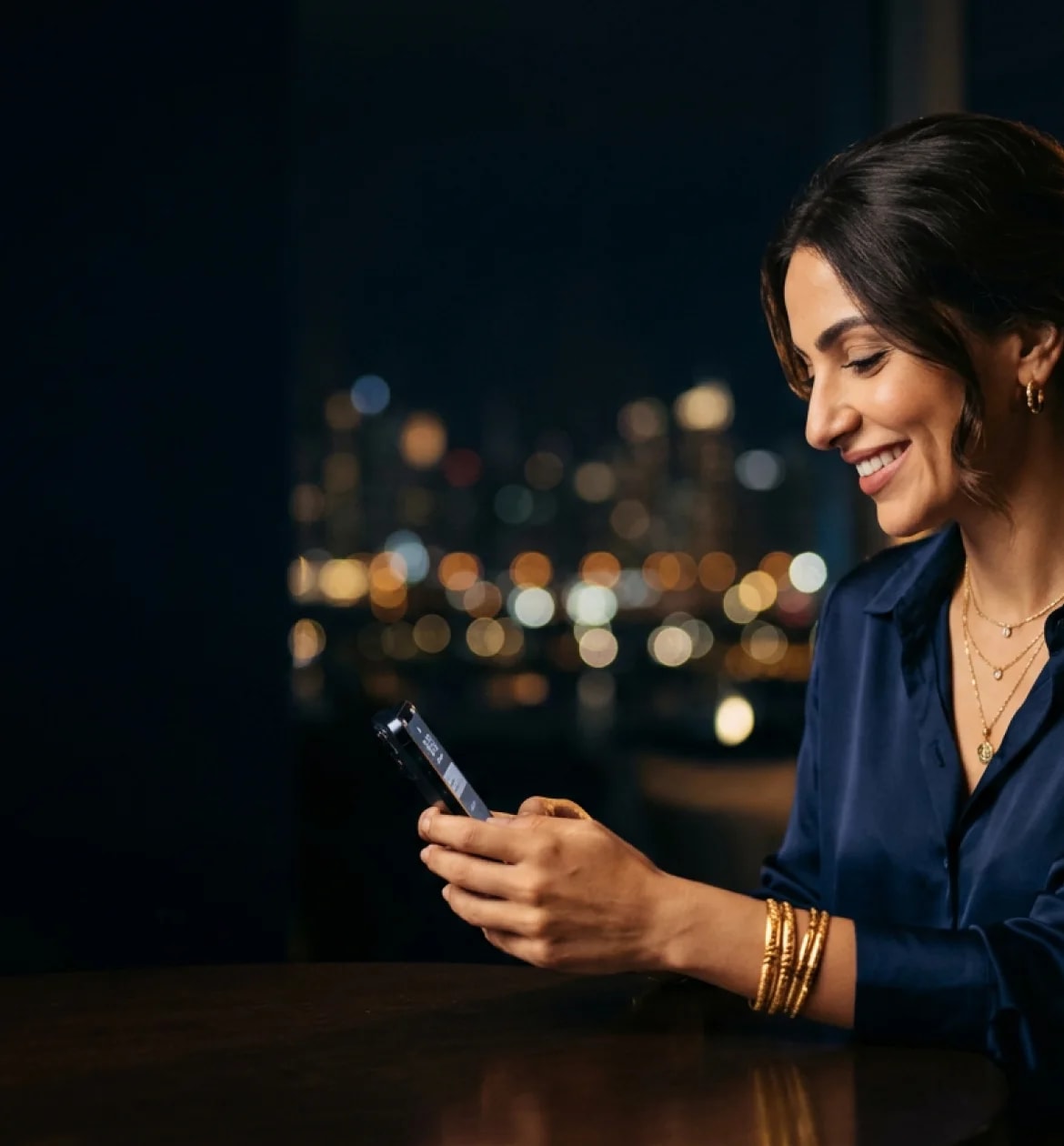 A woman in gold bangles smiling at her phone, city lights behind her